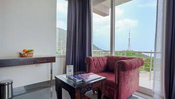 Een kamer met een rode fluweel fauteuil, een klein tafeltje met tijdschriften, een mand vol fruit en een uitzicht op een toren door grote ramen.
