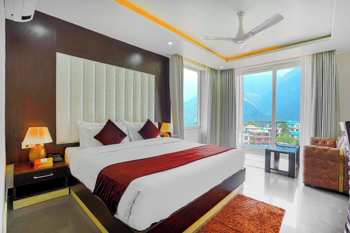 A hotel room with a large bed, a ceiling fan, a view of mountains, and a balcony with a chair.