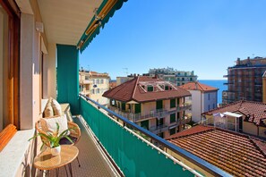 Un balcón con una silla de mimbre y una planta en maceta, con vistas a un pueblo costero de edificios con tejados de teja y un cielo azul despejado.