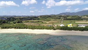 A coastal area with clear turquoise waters, sandy beach, and scattered buildings including a yellow house. - Ocean Villa Tokunoshima (Tokunoshima)