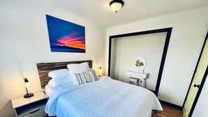 A bedroom with a bed, bedside tables, a nightstand, a dresser, a mirror, and a painting of a sunset.