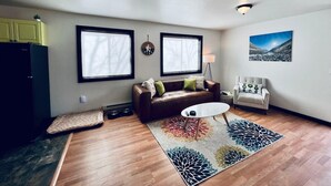 A living room with a brown leather sofa, a white armchair, a coffee table, and a colorful rug. There are two windows with blinds, a wall clock, and a framed picture on the wall.