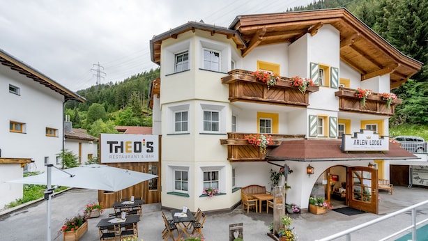 Un edificio a più piani con un ristorante chiamato "Theo's" e "Arlen Lodge" al piano terra. Sono presenti aree con posti a sedere all'aperto, tavoli, sedie e fioriere.
