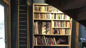 A room with a wooden bookshelf filled with books, a ladder leaning against the wall, and a wooden floor.