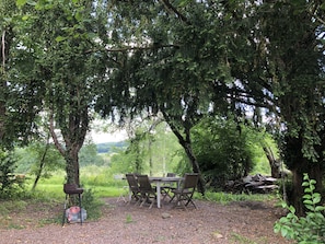 An outdoor dining area with a table and chairs under a large tree, surrounded by greenery and a view of the landscape.