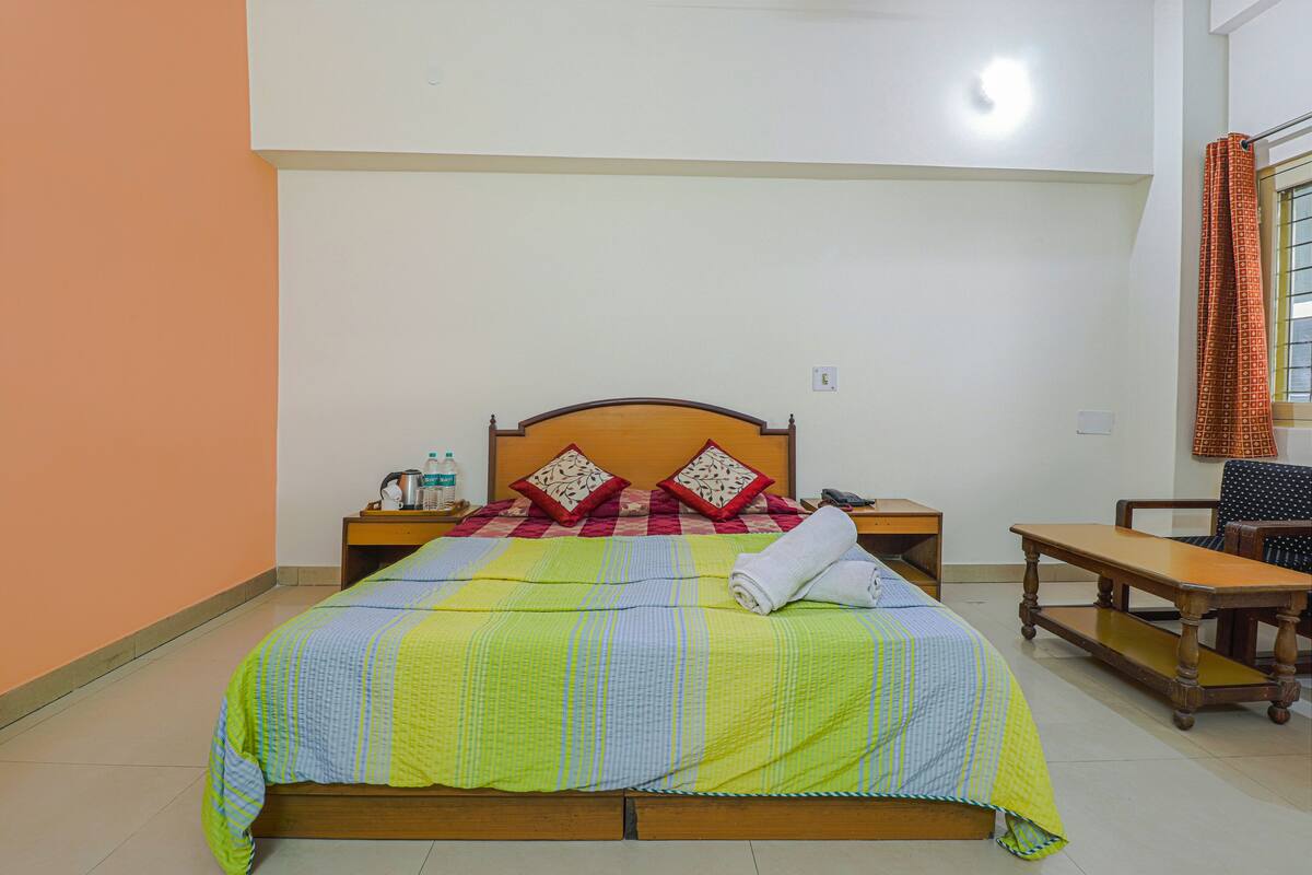 A neatly made bed with a striped bedspread, two pillows, a wooden headboard, a nightstand with a bottle and a cup, and a chair with a table beside the bed.
