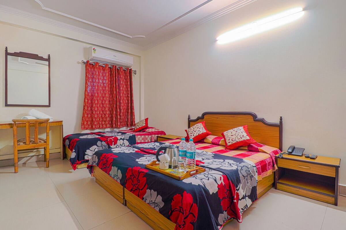 A double bed with floral bedding, a bedside table with a lamp, a desk with a chair, and a window with red curtains.