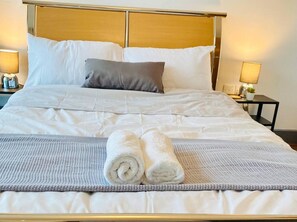 A neatly made bed with white linens, a grey and white textured blanket, and two rolled white towels.
