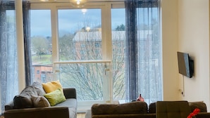 Ein geräumiges Wohnzimmer mit großem Fenster, einer Couch, einem Sessel und einem wandmontierten Flachbildfernseher.