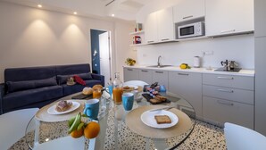 Une cuisine moderne avec des meubles blancs, un four à micro-ondes et un évier. On y trouve une table à manger avec des assiettes, des tasses et un verre de jus d’orange. Un canapé de couleur foncée se trouve au fond de la pièce.