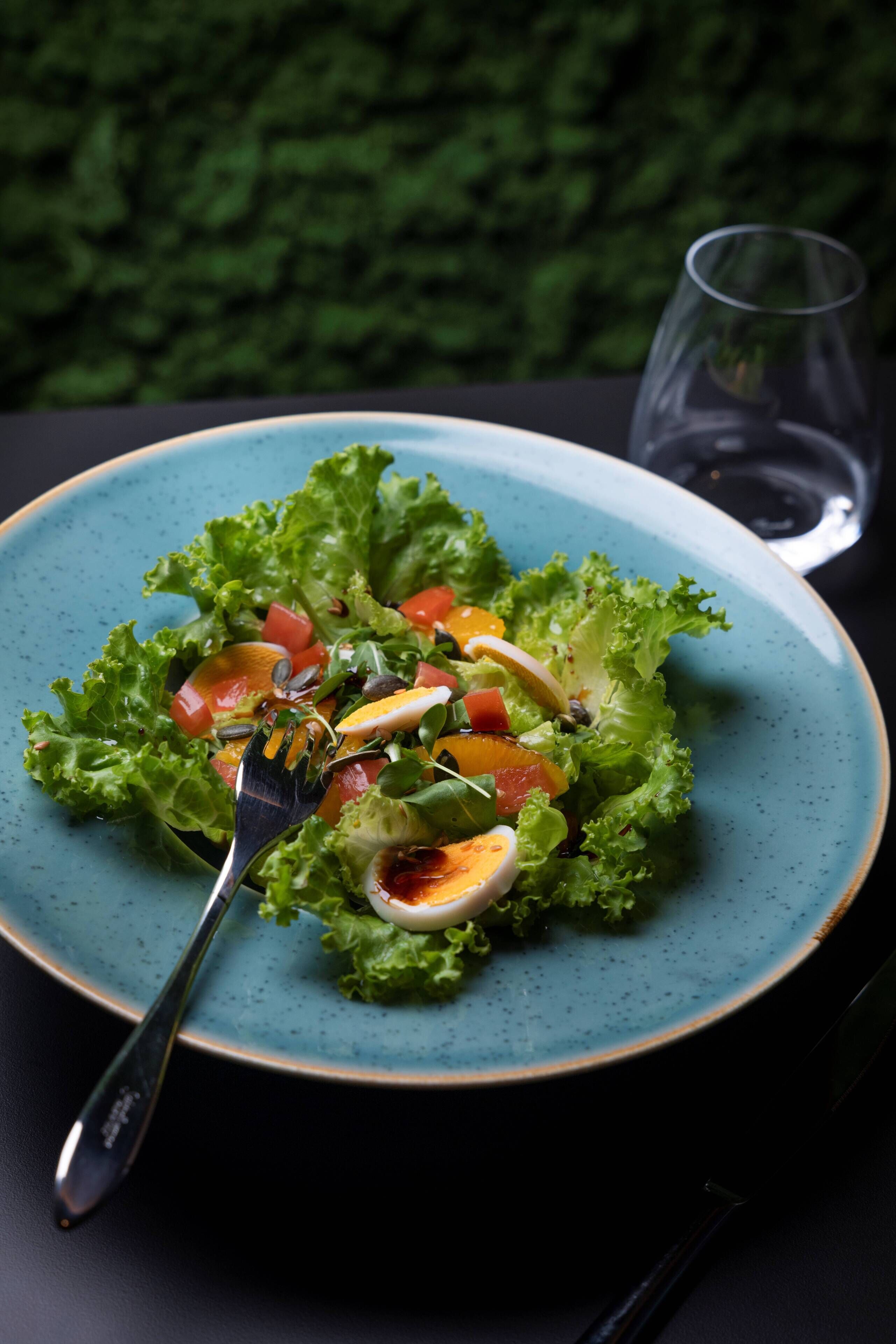 A plate of salad with a fork on a blue plate, a glass of water, and a dark background.
