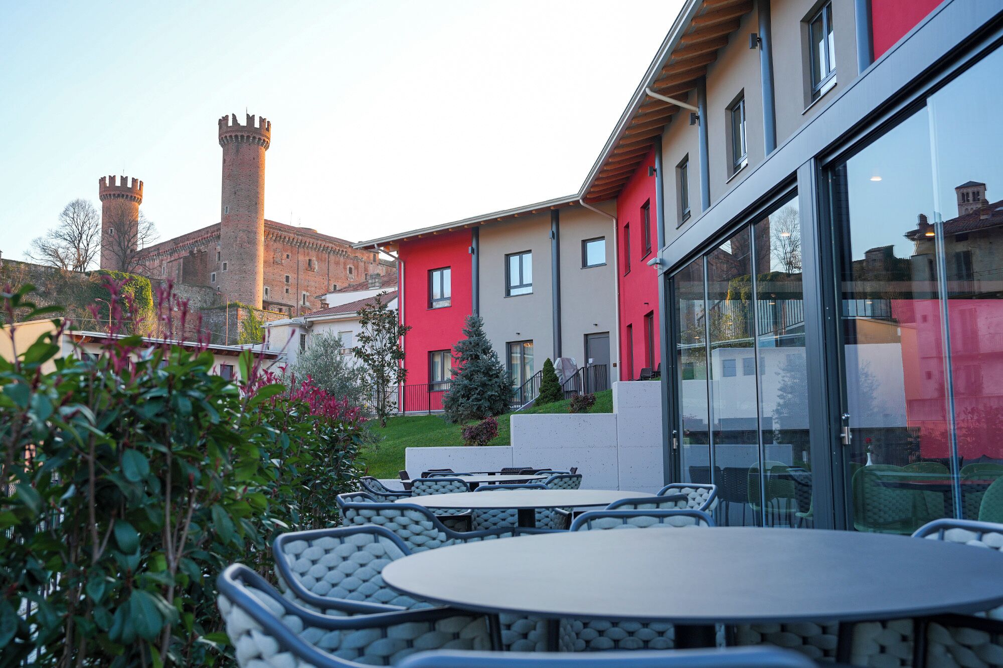 Un patio con mobiliario exterior, un edificio rojo y una torre histórica al fondo.