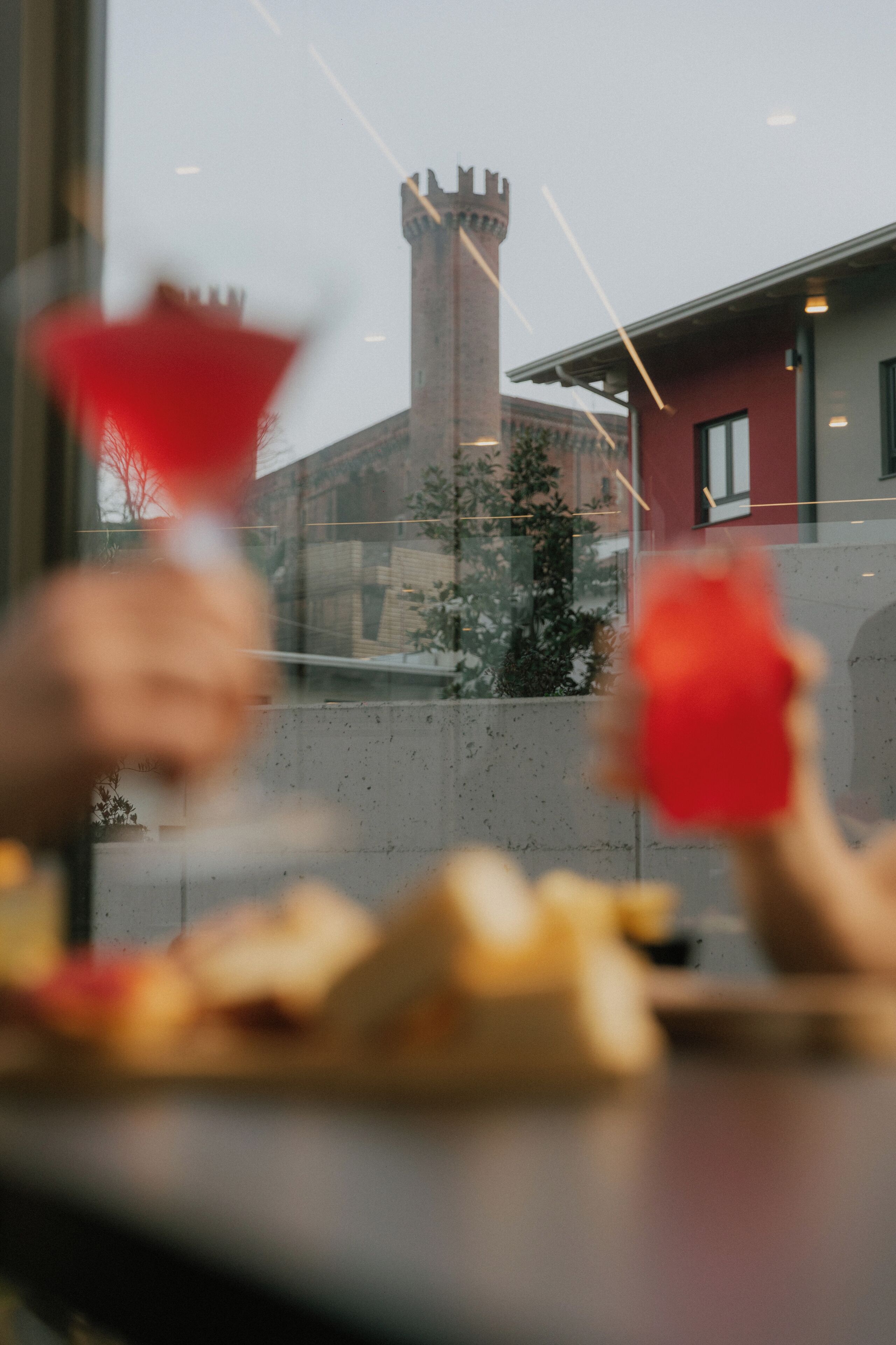Vista a través de una ventana que muestra una torre al fondo, una mesa con comida y un objeto rojo en primer plano.