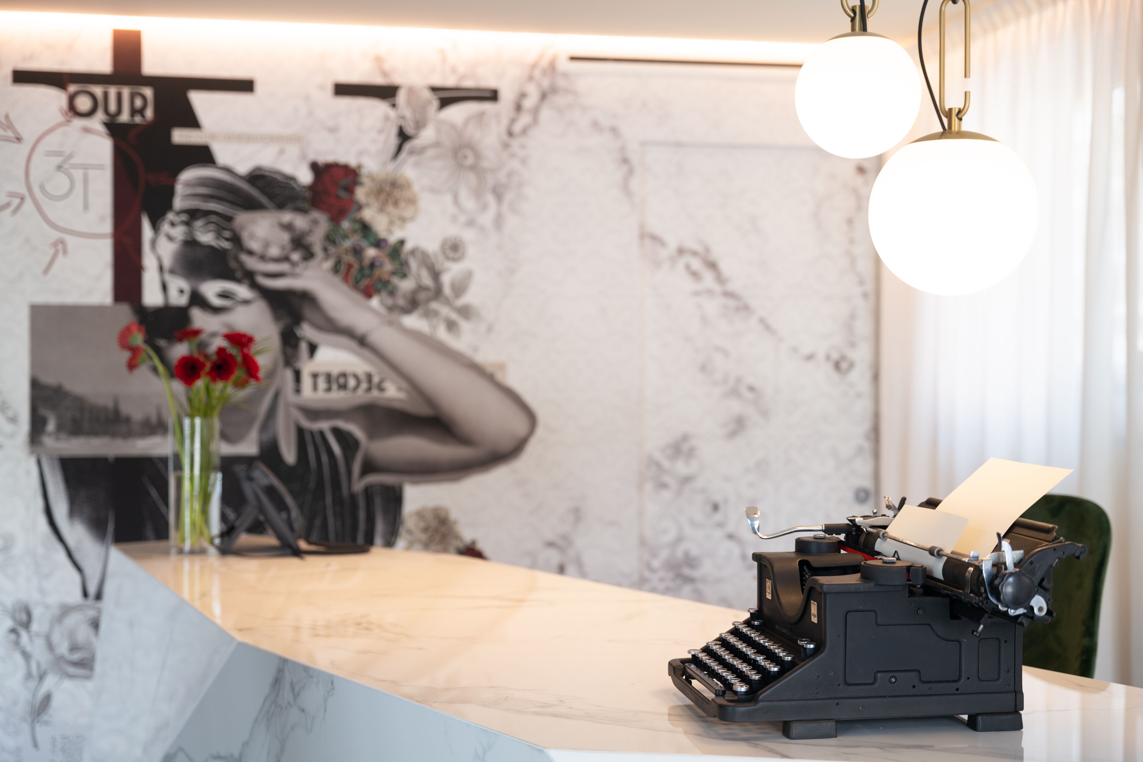 A vintage typewriter on a desk with a vase of red flowers, a wall with a floral mural, and a sign that reads "OUR".