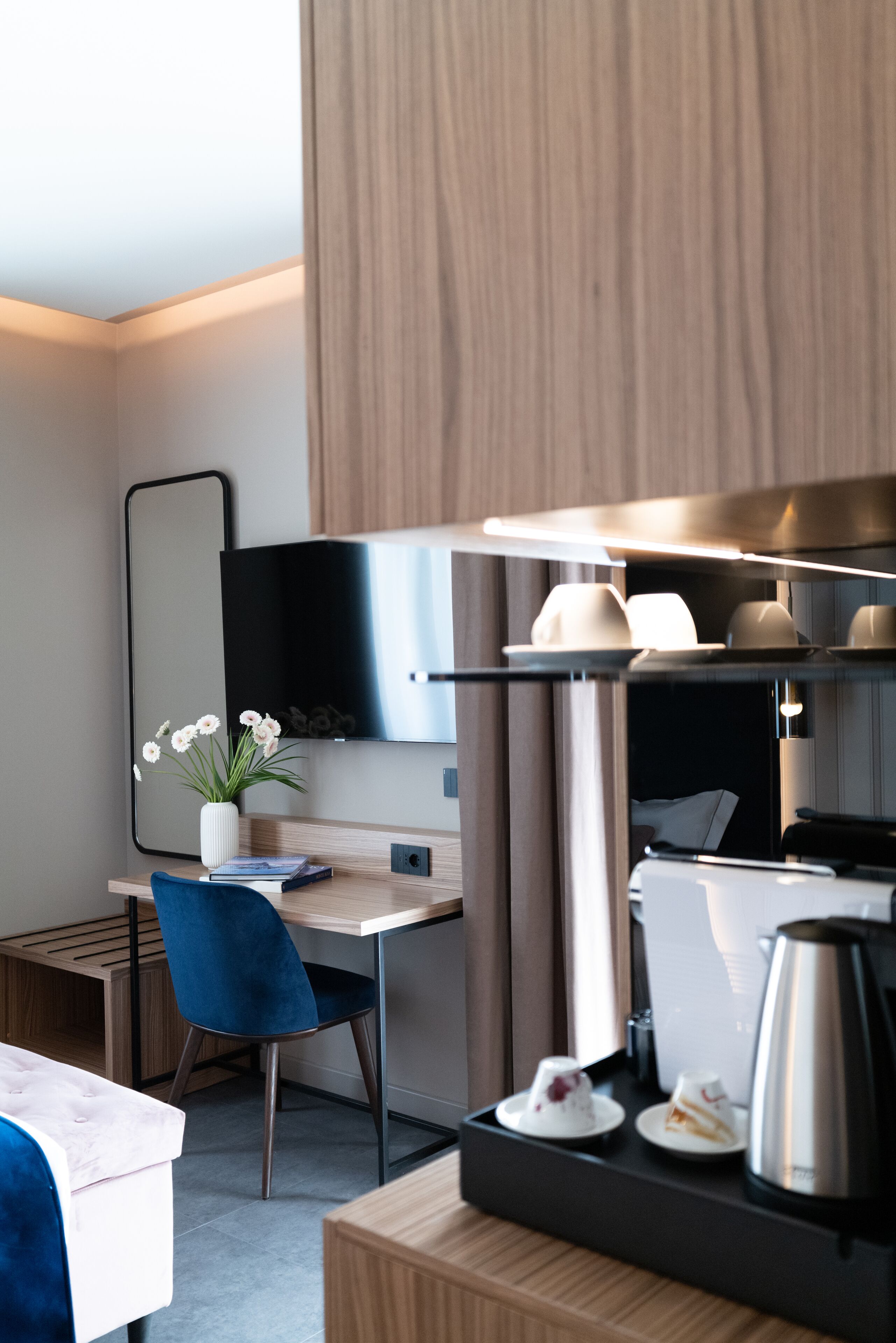 Ein modernes Hotelzimmer mit einem Holztisch, einem blauen Sessel, einem Flachbildfernseher, einer Kaffeemaschine und einer kleinen Pflanze.