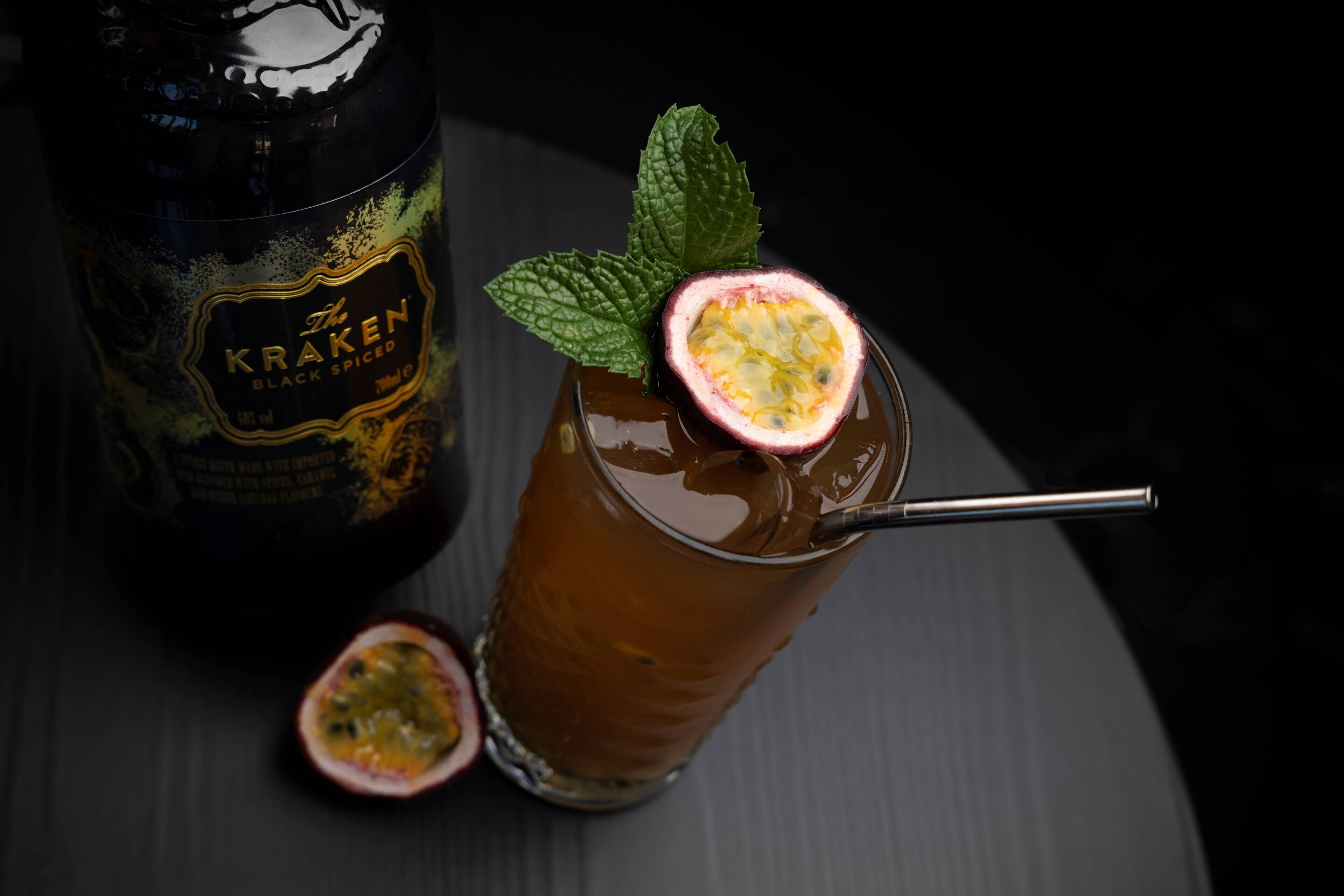 A glass of a beverage with a passion fruit garnish, a bottle labeled "The Kraken Black Spiced," and a mint leaf.