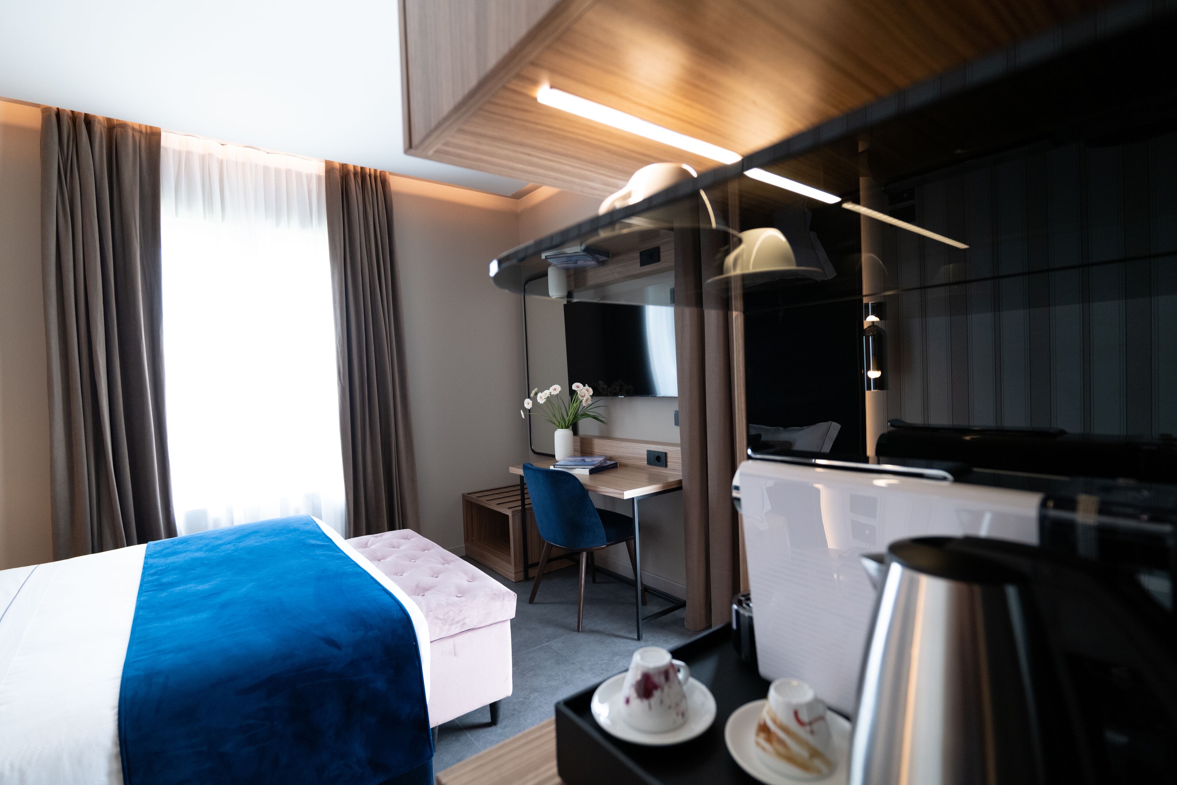 Una habitación de hotel moderna con una cama, un escritorio, una silla, una mesita con una cafetera y una ventana con cortinas.
