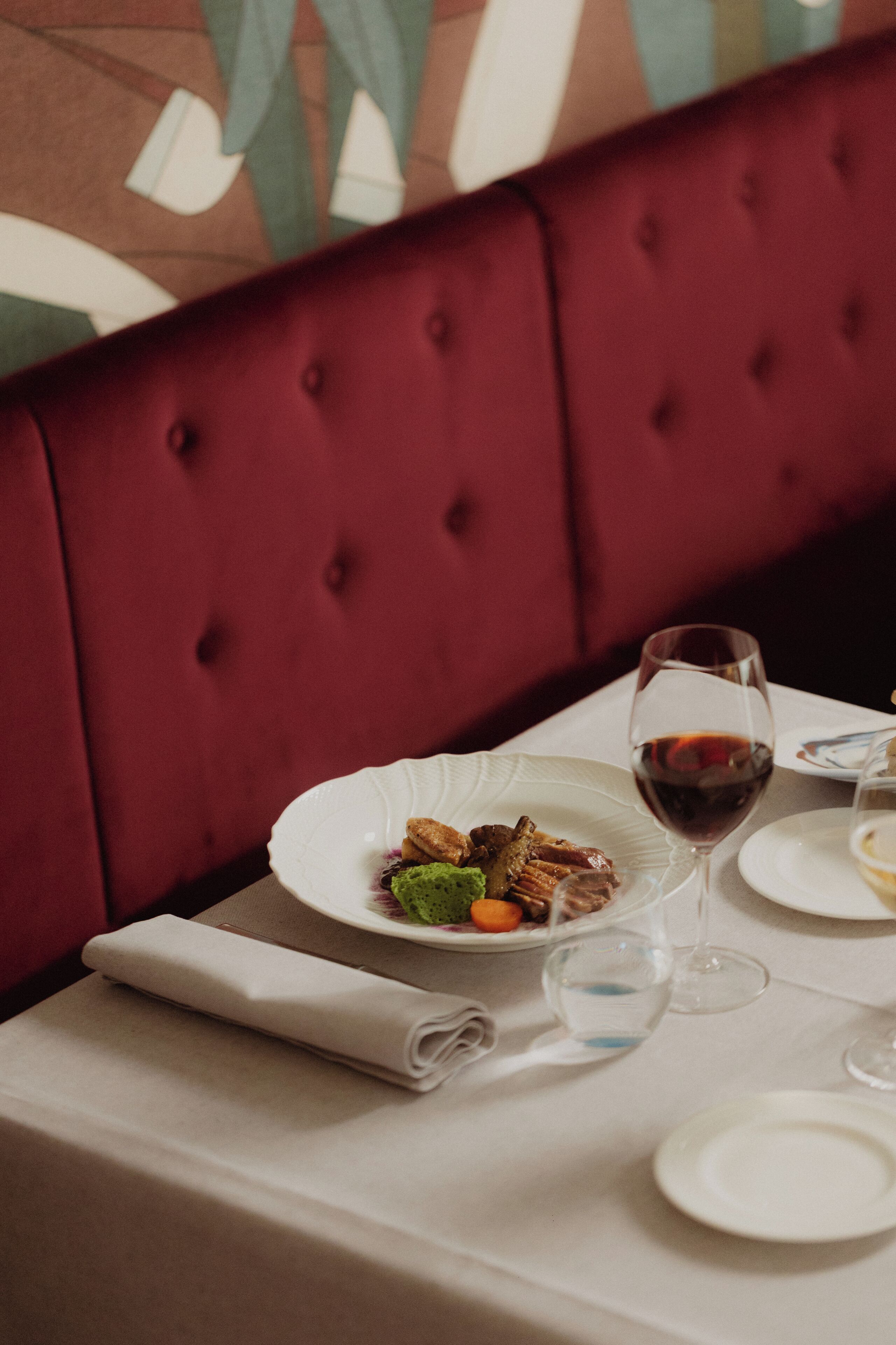 Una mesa de comedor con mantel blanco, servilleta doblada, plato de comida, copa de vino tinto y vaso de agua.