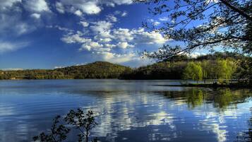 Un llac tranquil amb reflexos d'arbres i núvols, envoltat de turons i un cel serè.