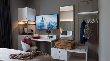 A modern hotel room with a bed, a desk with a flat-screen TV, a vanity with a mirror, a wardrobe, and a large window with a cityscape view.