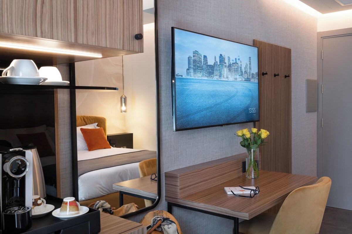 Una habitación de hotel moderna con cama, mesita de noche, escritorio con silla, televisor mostrando un paisaje urbano y un florero con flores.