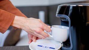 Een hand plaatst een witte kop op een schotel, naast een koffiezetapparaat.