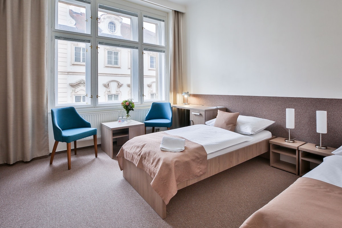 Habitación de hotel con cama, dos sillas, un escritorio, una lámpara y ventana con vistas a edificios.