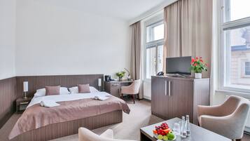 A hotel room with a bed, desk, chair, TV, and a table with fruit and bottles.