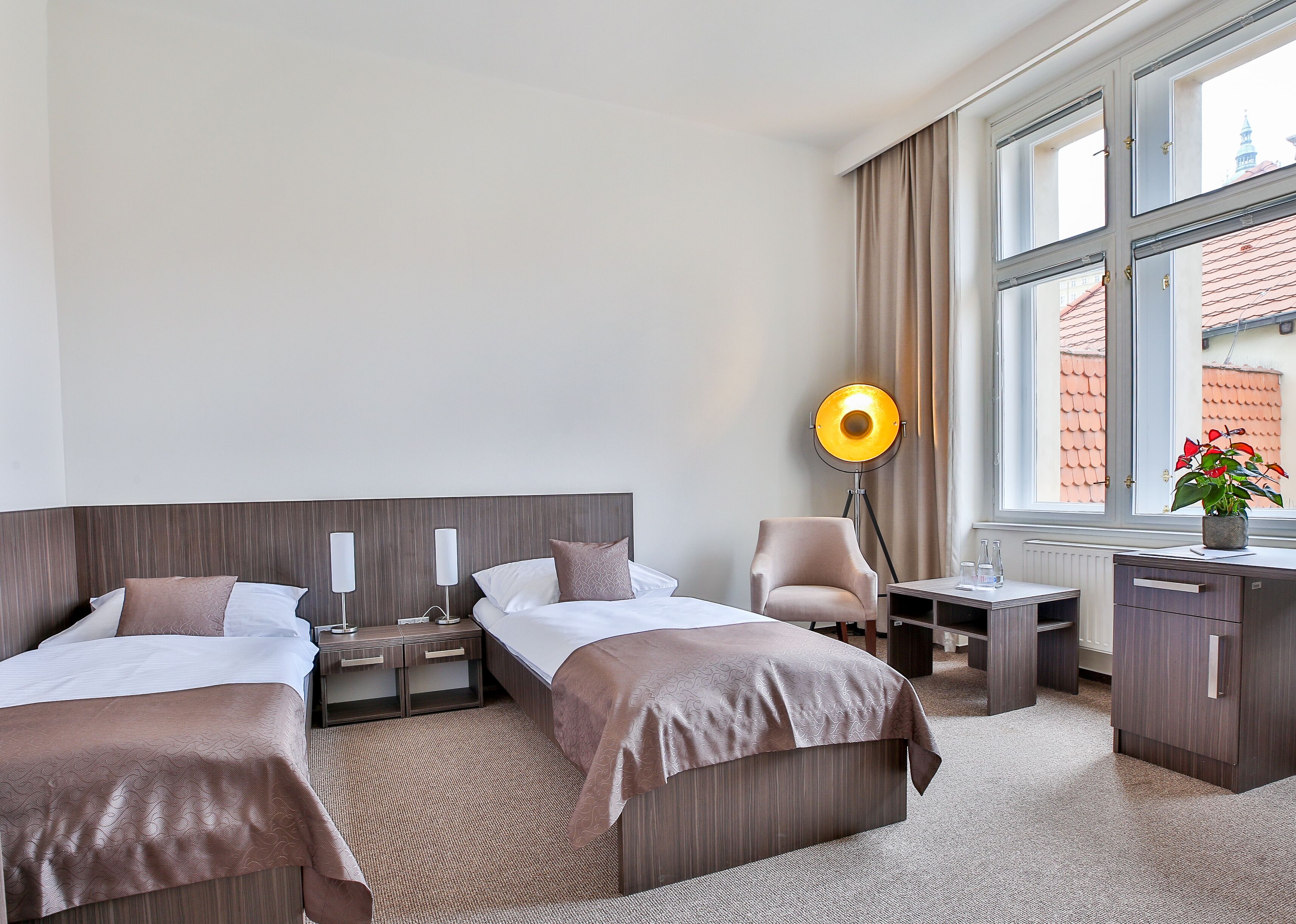 A hotel room with two beds, a desk, a chair, a floor lamp, and a view of buildings through large windows.