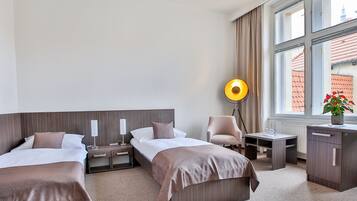 A hotel room with two beds, a desk, a chair, a floor lamp, and a view of buildings through large windows.