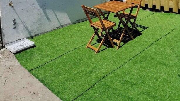 Een huis met een witte gevel, een dak met rode tegels, een houten omheining en een klein terras met kunstgras, een houten tafel en stoelen.