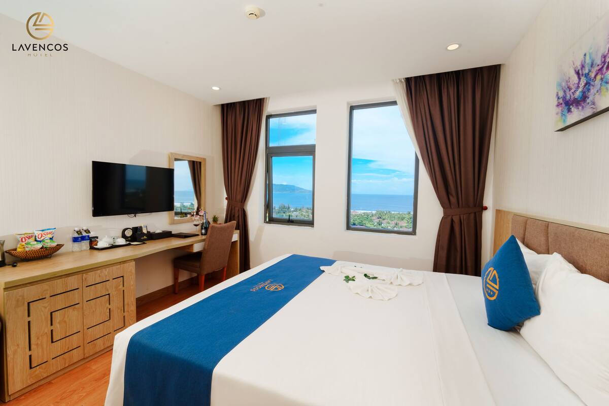 A hotel room with a bed, a desk, a television, and a view of the sea.