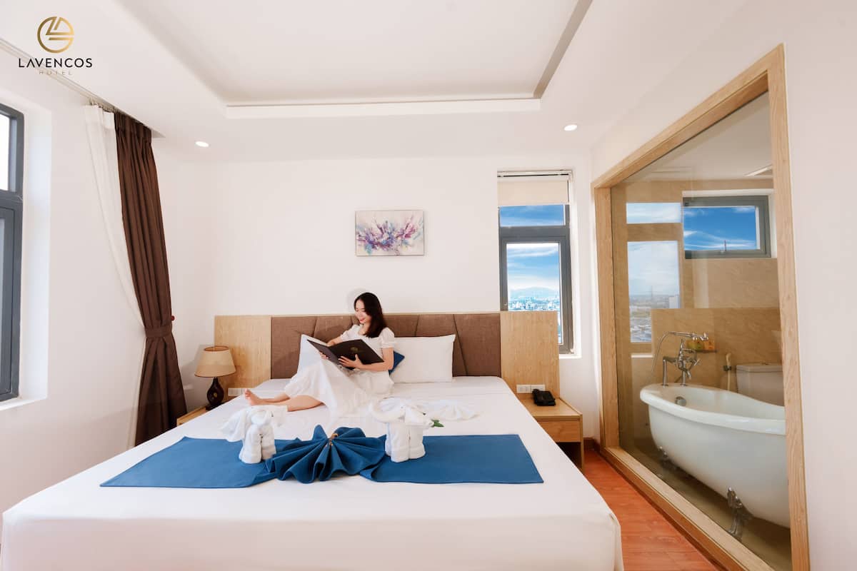 A hotel room with a large bed, a reading area, a bathroom with a bathtub, and a city view.