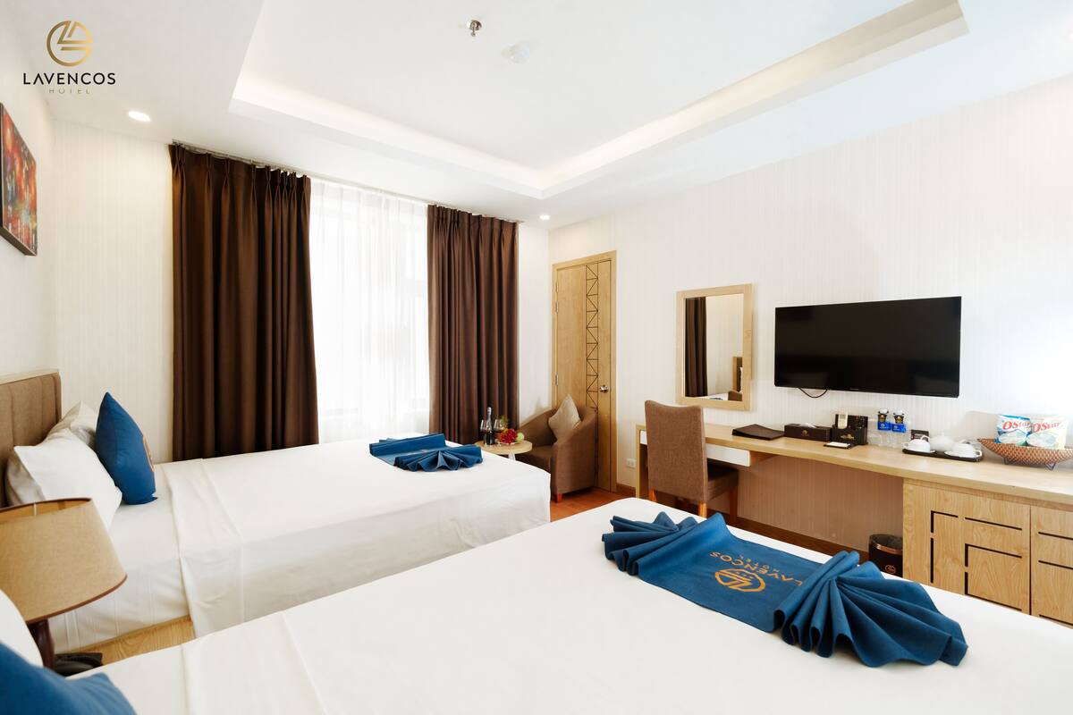 A hotel room with two beds, a desk, a chair, a television, and a large window with curtains.