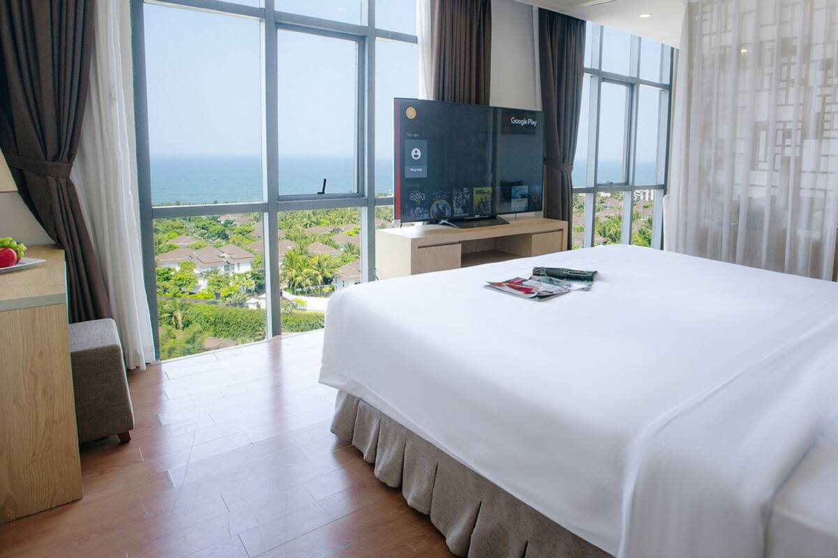 A hotel room with a large bed, a TV on a stand, and a view of the ocean and greenery.