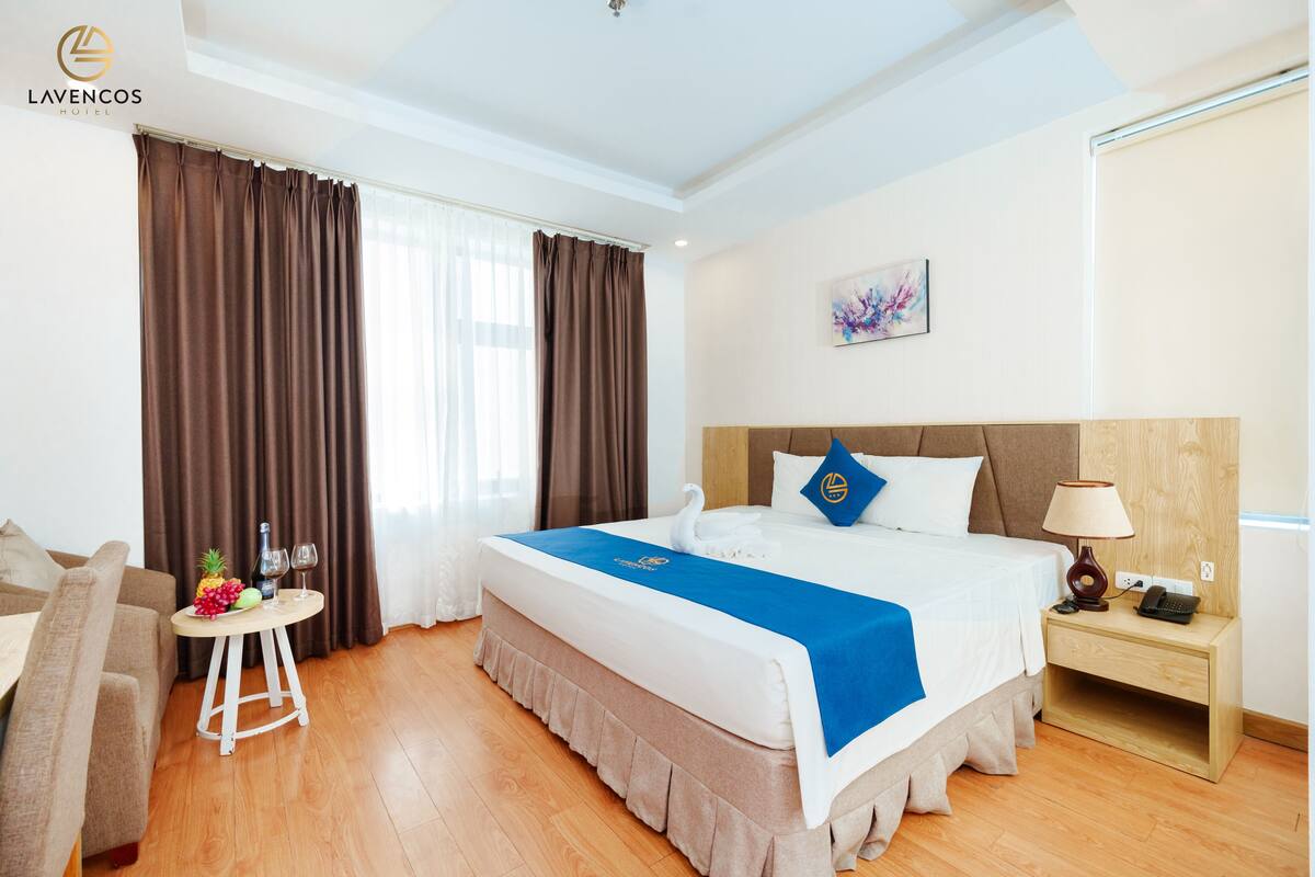 A hotel room with a large bed, a small table with a fruit bowl, a lamp, and a nightstand.