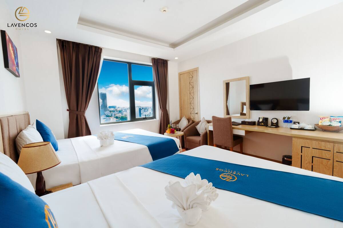 A hotel room with two beds, a desk, a TV, and a window with a city view.