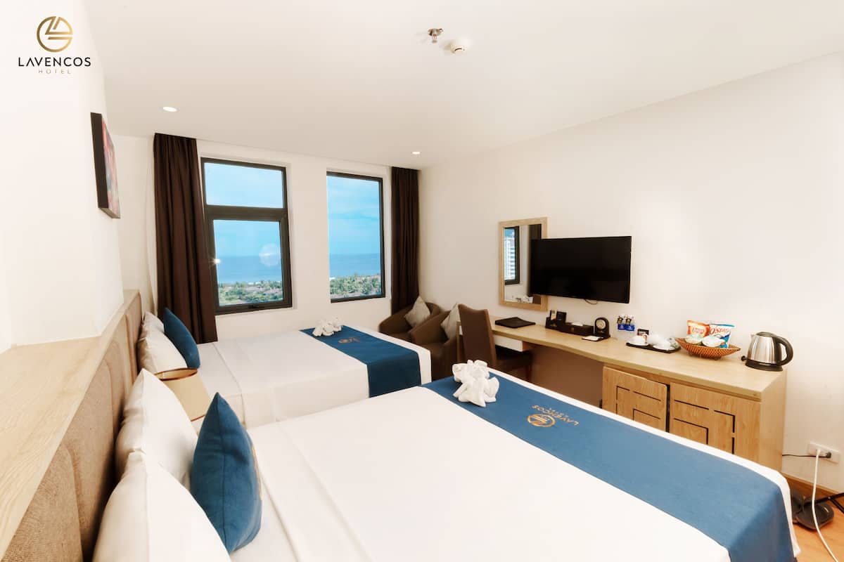 A hotel room with two beds, a desk, a TV, and a view of the sea.