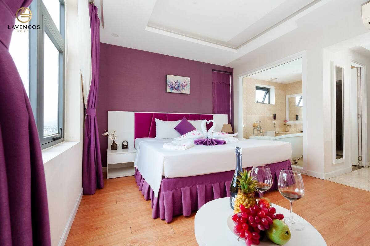 A modern hotel room with a large bed, purple bedding, a round table with fruit and wine, and a bathroom visible through an open door.