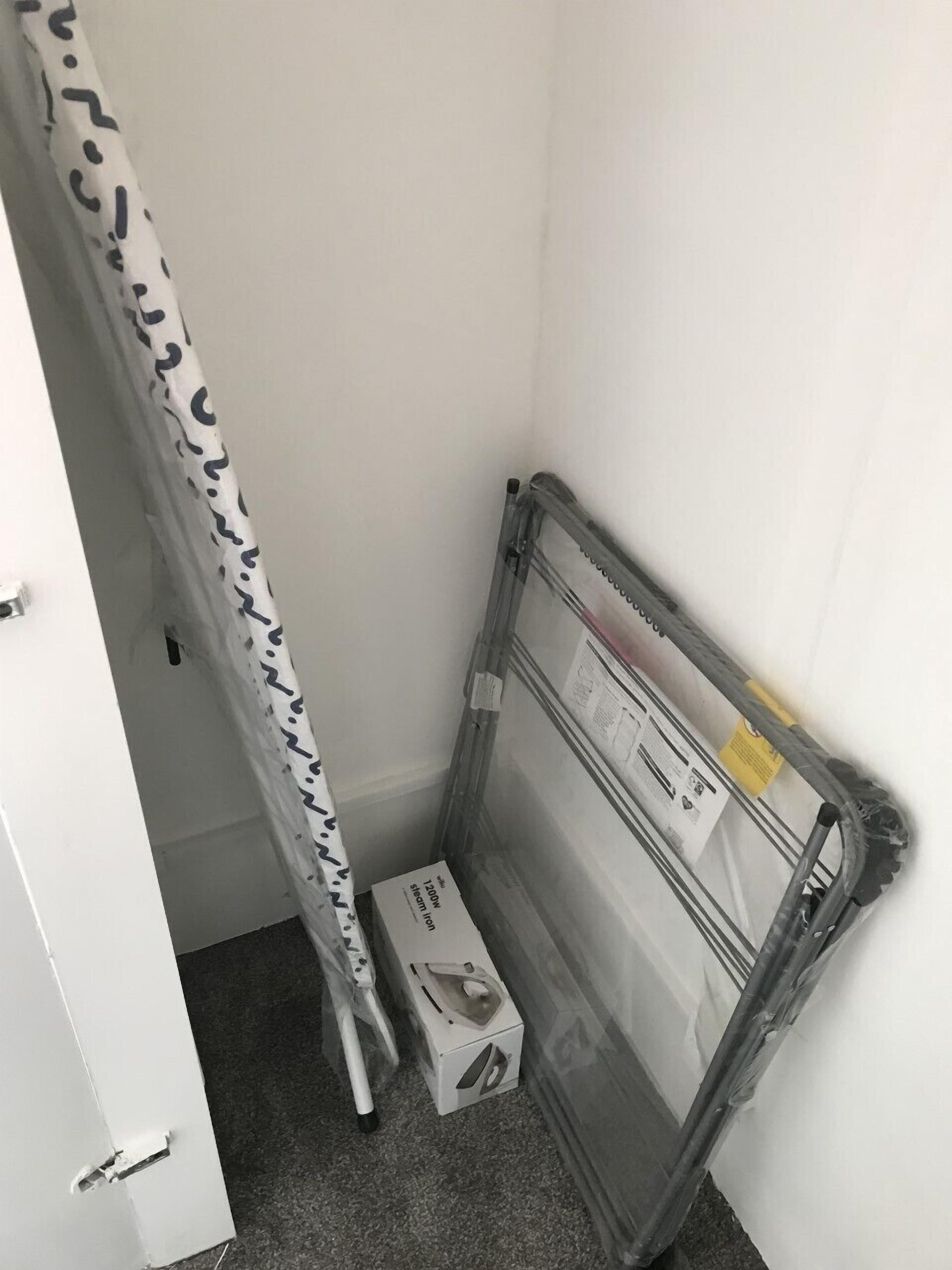Esquina de una habitación con una puerta blanca, una cortina estampada, una escalera metálica y una caja etiquetada como '1200W steam iron'.