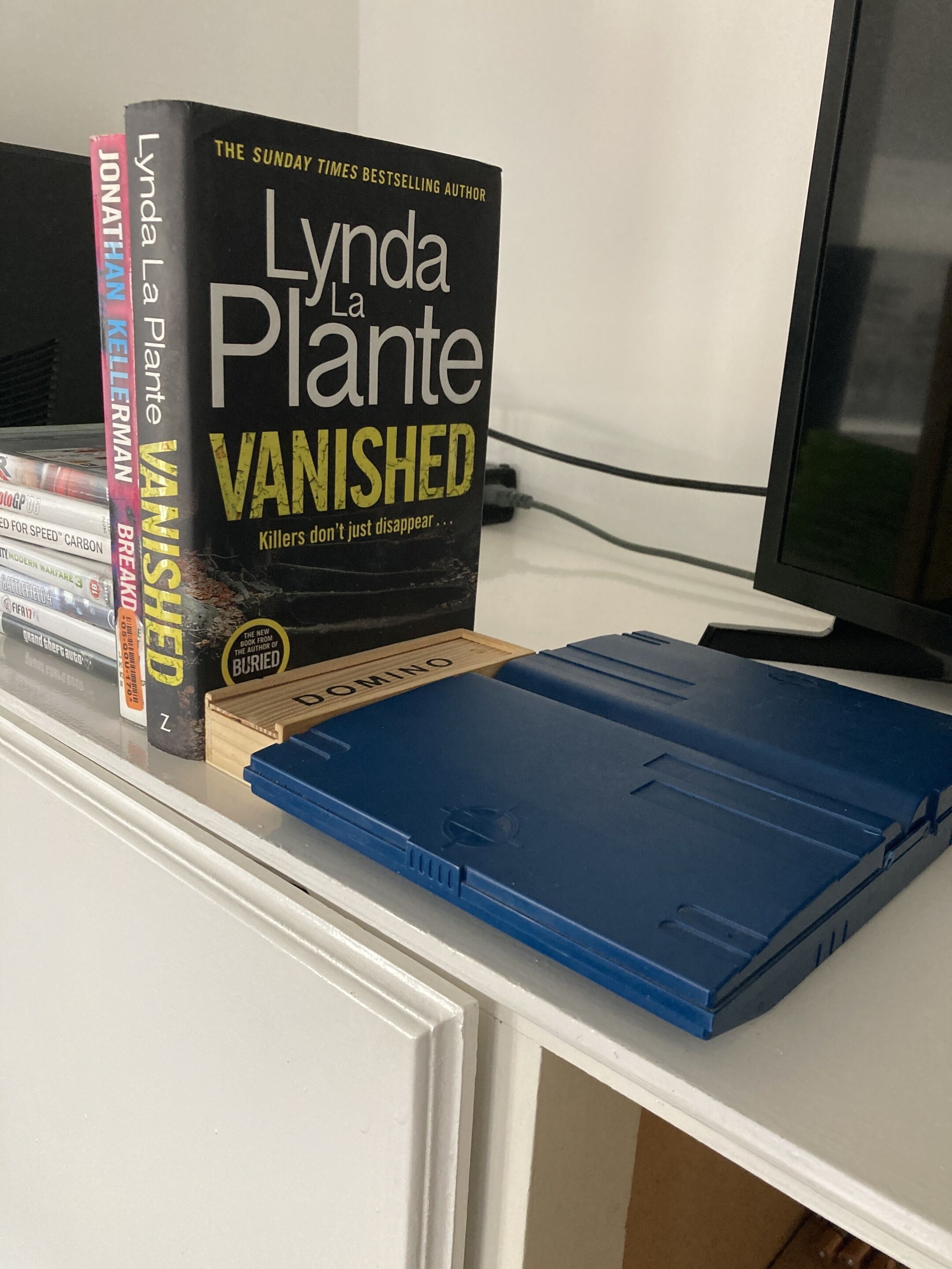 Een boek met de titel 'Vanished' van Lynda LaPlante op een witte plank, naast een blauw boek en een houten doos.