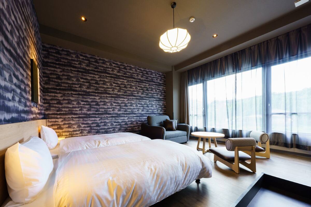 Een moderne hotelkamer met een groot bed, een bank, een tafeltje en uitzicht op het landschap door grote ramen.