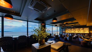 Un intérieur moderne avec une grande fenêtre donnant sur la mer, des espaces aménagés pour s’asseoir et une plante décorative placée au centre.