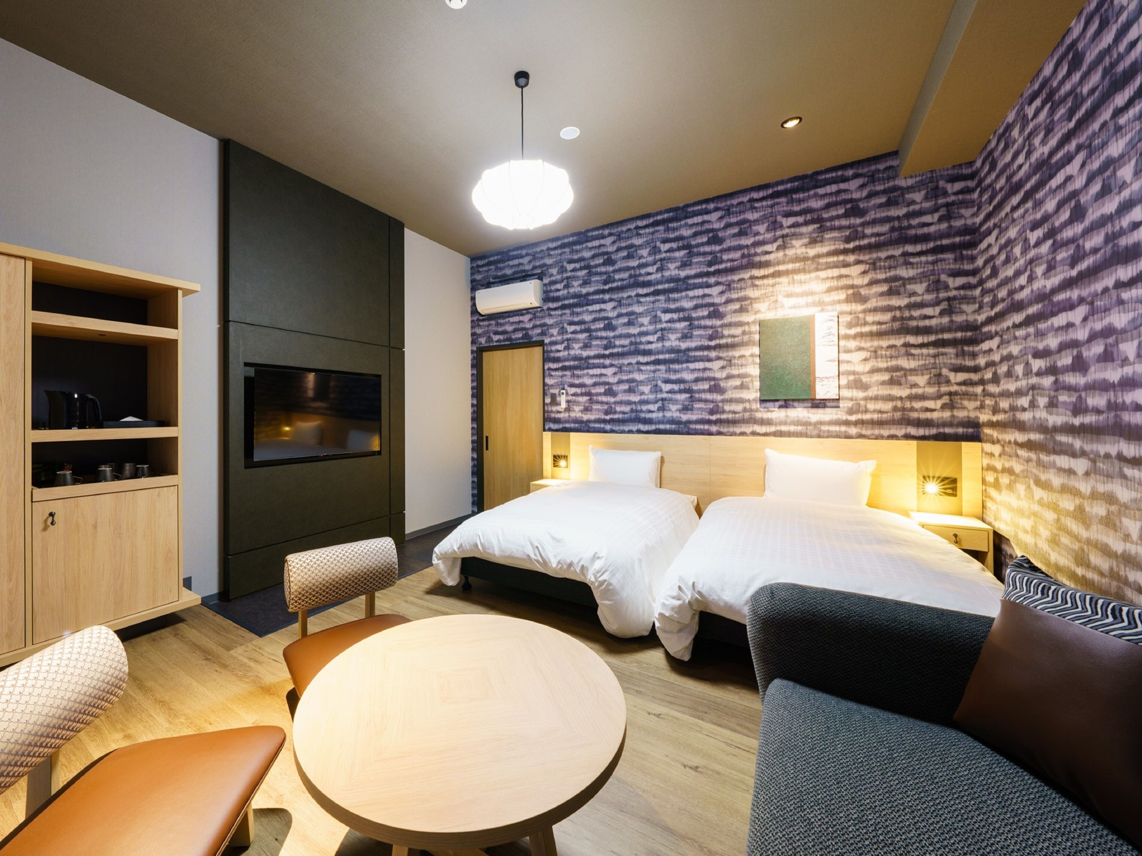 A modern hotel room with two beds, a dining table, a sofa, a TV, and a built-in kitchen area.