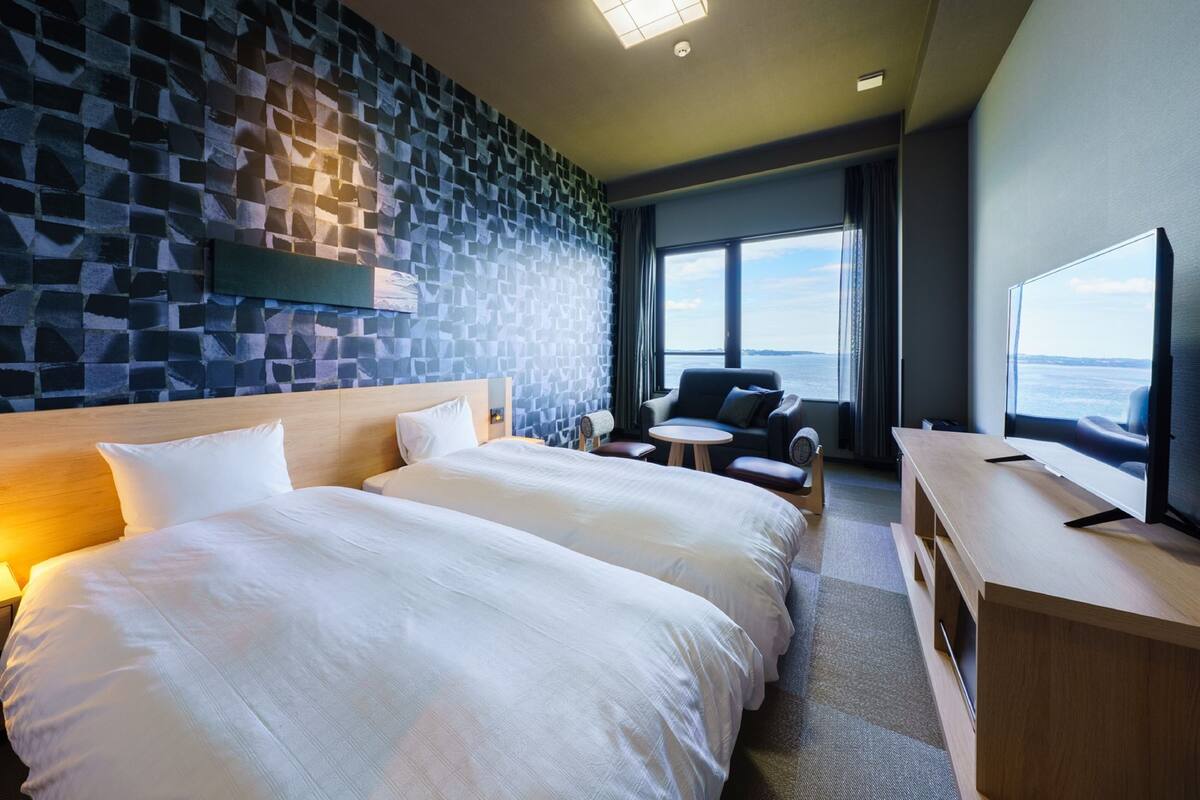 Een hotelkamer met twee tweepersoonsbedden, een zithoek met uitzicht op zee, een flatscreen televisie en een wand met een stenen patroon.