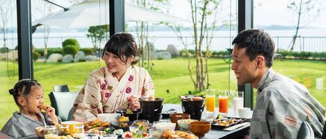 Una mesa de comedor con diversos platos y bebidas, ventanas amplias con vistas a un área verde y árboles, y una persona vestida con traje tradicional.