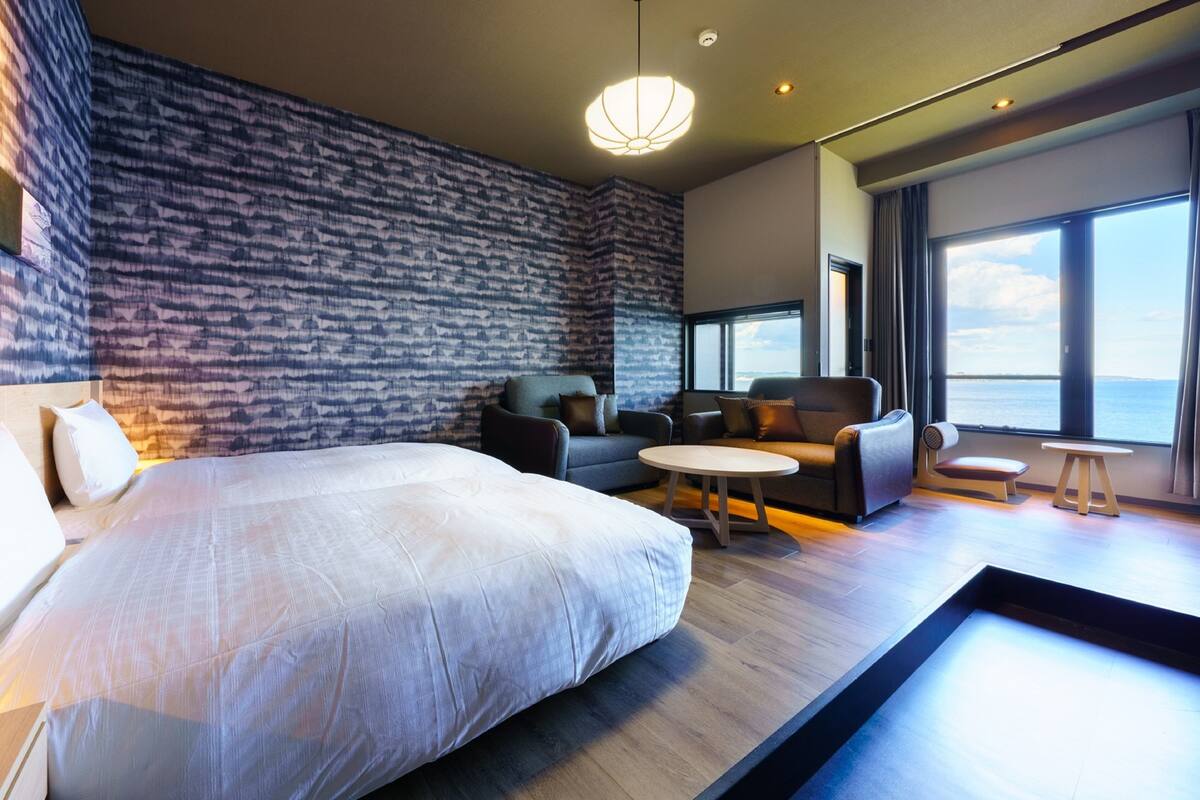 Een moderne hotelkamer met een groot bed, een zithoek met banken en een kleine tafel, een behang met een patroon, en een groot raam met uitzicht.