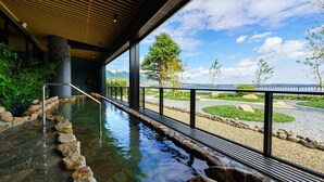 Un espace de sources chaudes naturelles, avec des pierres, un plafond en bois, une rambarde en métal et une vue sur un jardin aménagé et des montagnes.