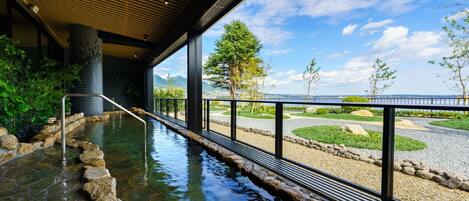 A stone hot spring area with a wooden ceiling, a metal railing, and a view of a landscaped garden and mountains.