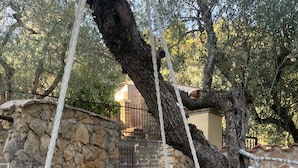 Une chaise suspendue en macramé blanc, agrémentée d’un coussin, est accrochée à un arbre dans un jardin dont les murs et l’escalier sont en pierre.